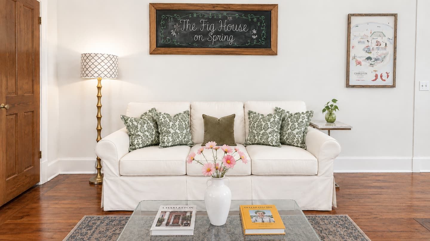 Ein Wohnzimmer mit einem weißen Sofa, gemusterten Kissen, einer Blumenvase, einer Lampe und einem gerahmten Bild an der Wand.