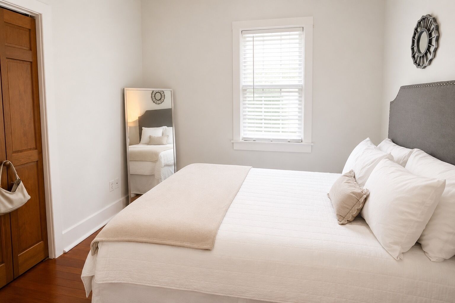 A bedroom with a bed, a window with blinds, a wooden door, and a mirror on the wall.