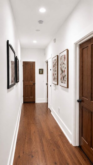A hallway with wooden flooring, white walls, and doors. There are framed pictures on the walls.