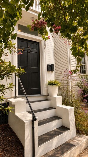 A white house with a black front door, a set of concrete steps, and a potted plant on the landing. - The Fig House on Spring · Walk to King · 3BR, Sleeps 8 (Charleston)