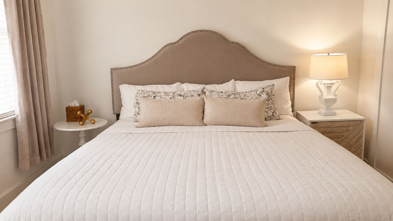 A neatly made bed with a headboard, a bedside table with a lamp, and a window with curtains.