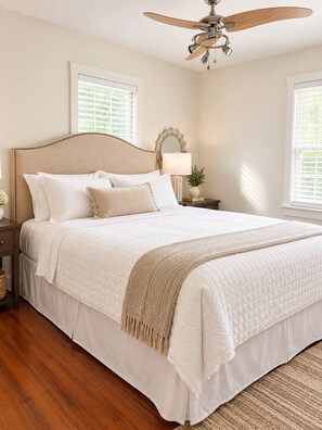 A bedroom with a large bed, a ceiling fan, two windows with blinds, a bedside table with a lamp, and a nightstand with a vase. - The Fig House on Spring · Walk to King · 3BR, Sleeps 8 (Charleston)