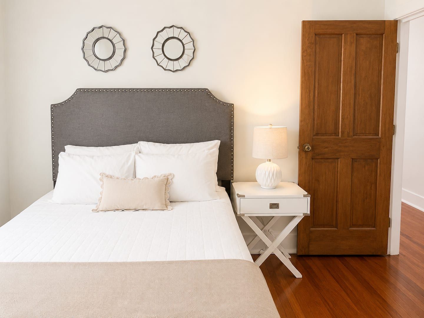 A neatly made bed with white bedding, a grey upholstered headboard, and a bedside table with a lamp.