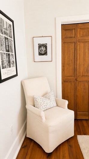A white armchair with a patterned pillow, a framed black and white photograph, and a wooden door in a room with wooden flooring. - The Fig House on Spring · Walk to King · 3BR, Sleeps 8 (Charleston)