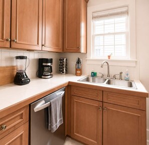 A kitchen with wooden cabinets, a coffee maker, a blender, a dishwasher, and a window with blinds.