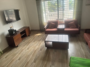 Una sala de estar con un conjunto de sofá, una mesita de centro, un televisor montado en la pared y un ventanal con cortinas.
