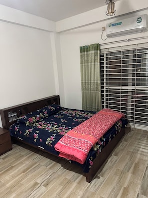 A bedroom with a bed, a nightstand, a window with blinds, and an air conditioning unit.
