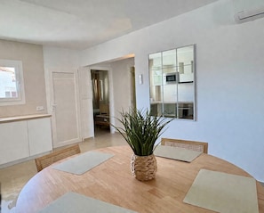 A modern kitchen with white cabinets, a wooden dining table with placemats, a potted plant, and a large mirror reflecting the kitchen. - Brisa del Mar 54 (Los Delfines)
