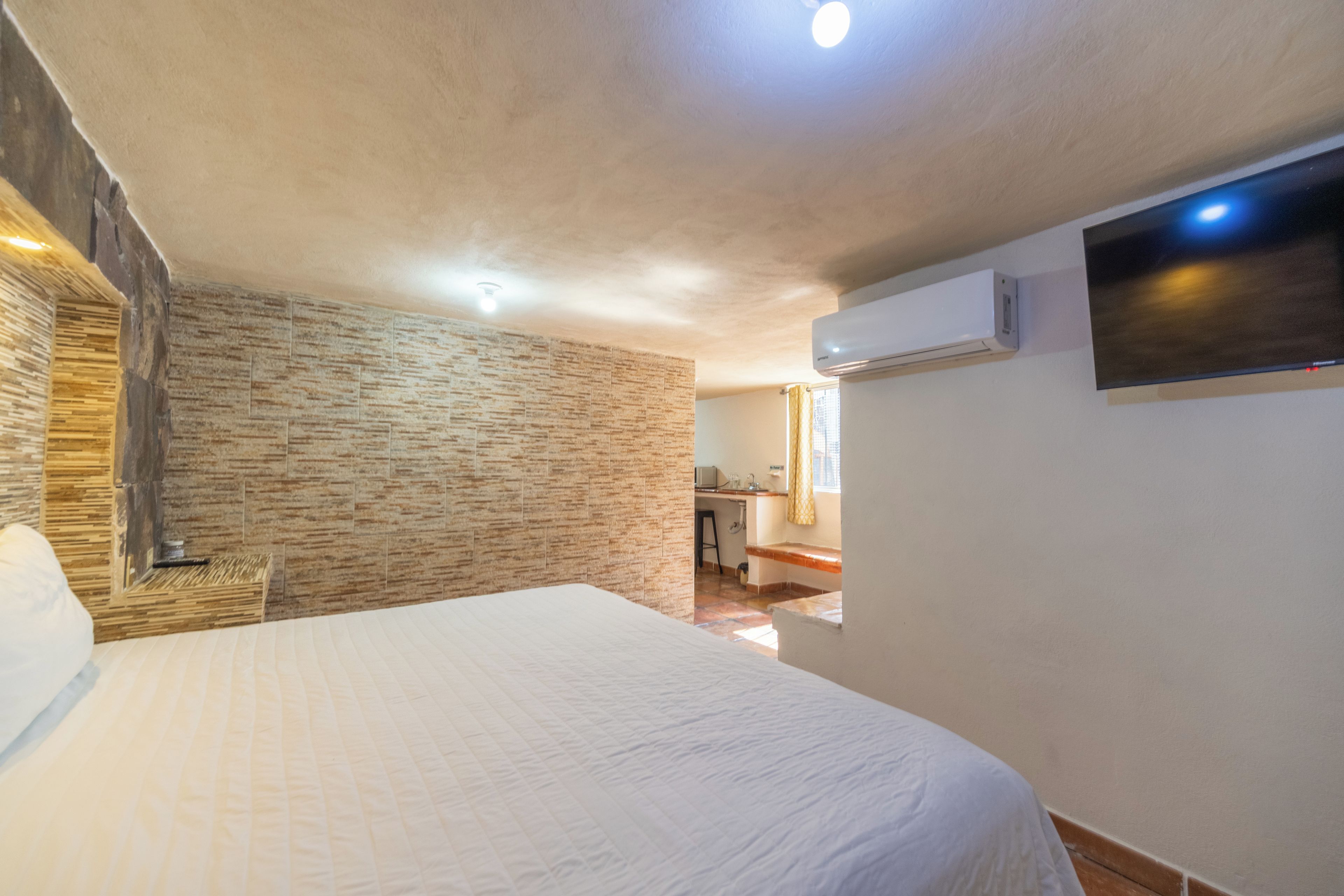 A bedroom with a stone wall, a bed, an air conditioning unit, and a view into the kitchen.