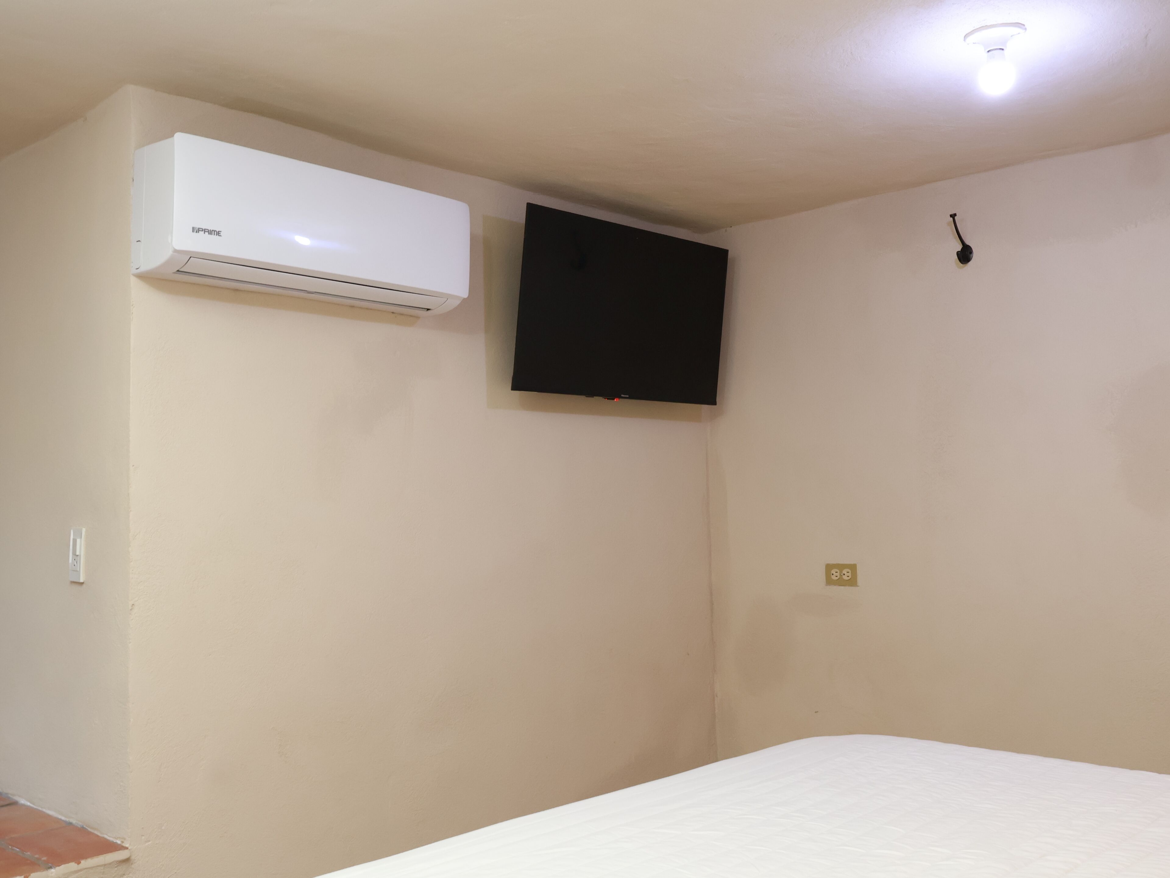 A room with a bed, an air conditioner, a wall-mounted TV, and a light fixture.
