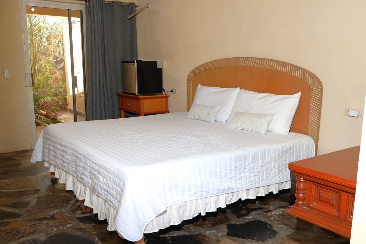 A bedroom with a large bed, a wooden nightstand, a television on the nightstand, and a stone floor.