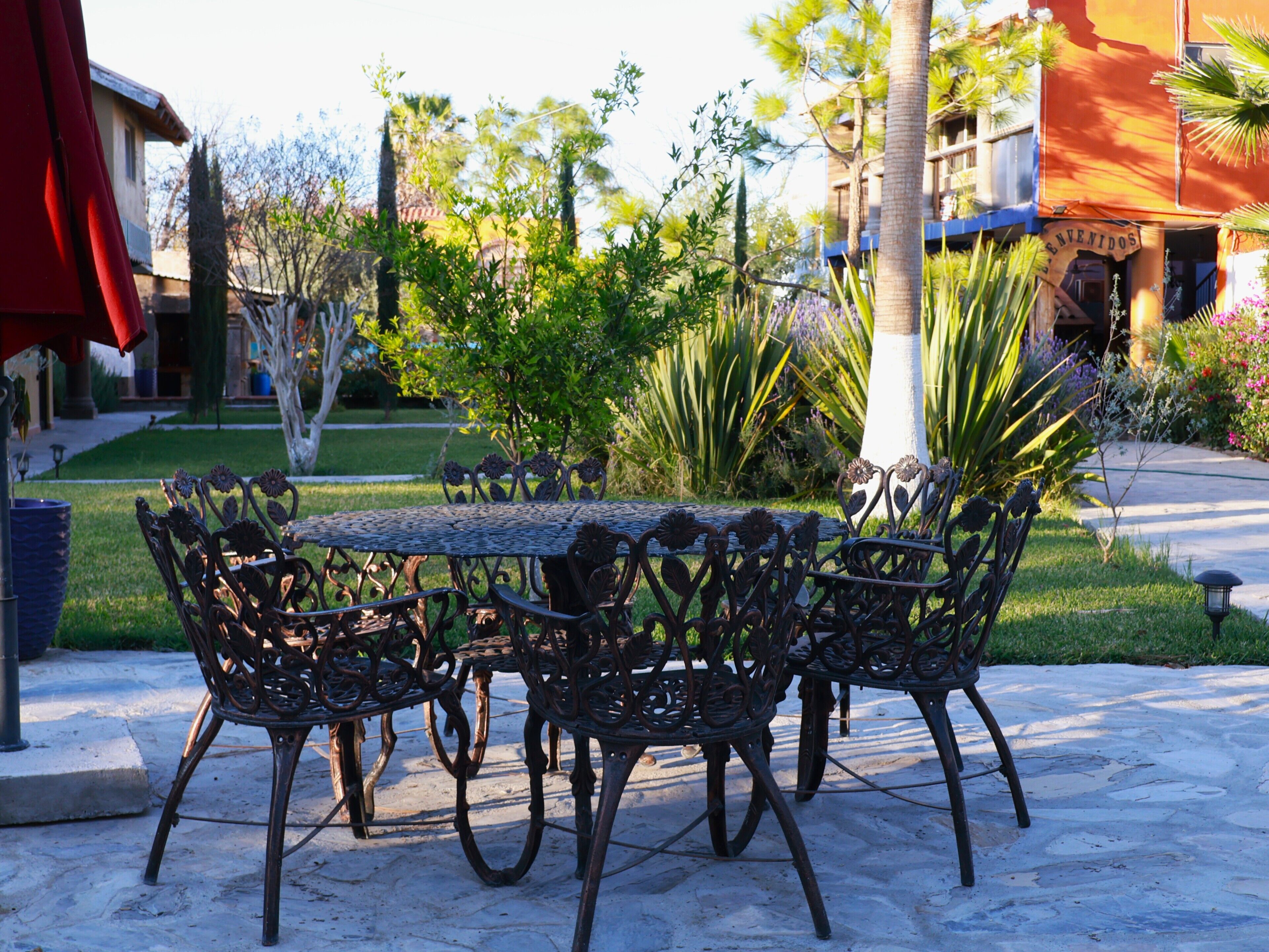 An outdoor patio with a round table and chairs, surrounded by greenery and a building with a sign.