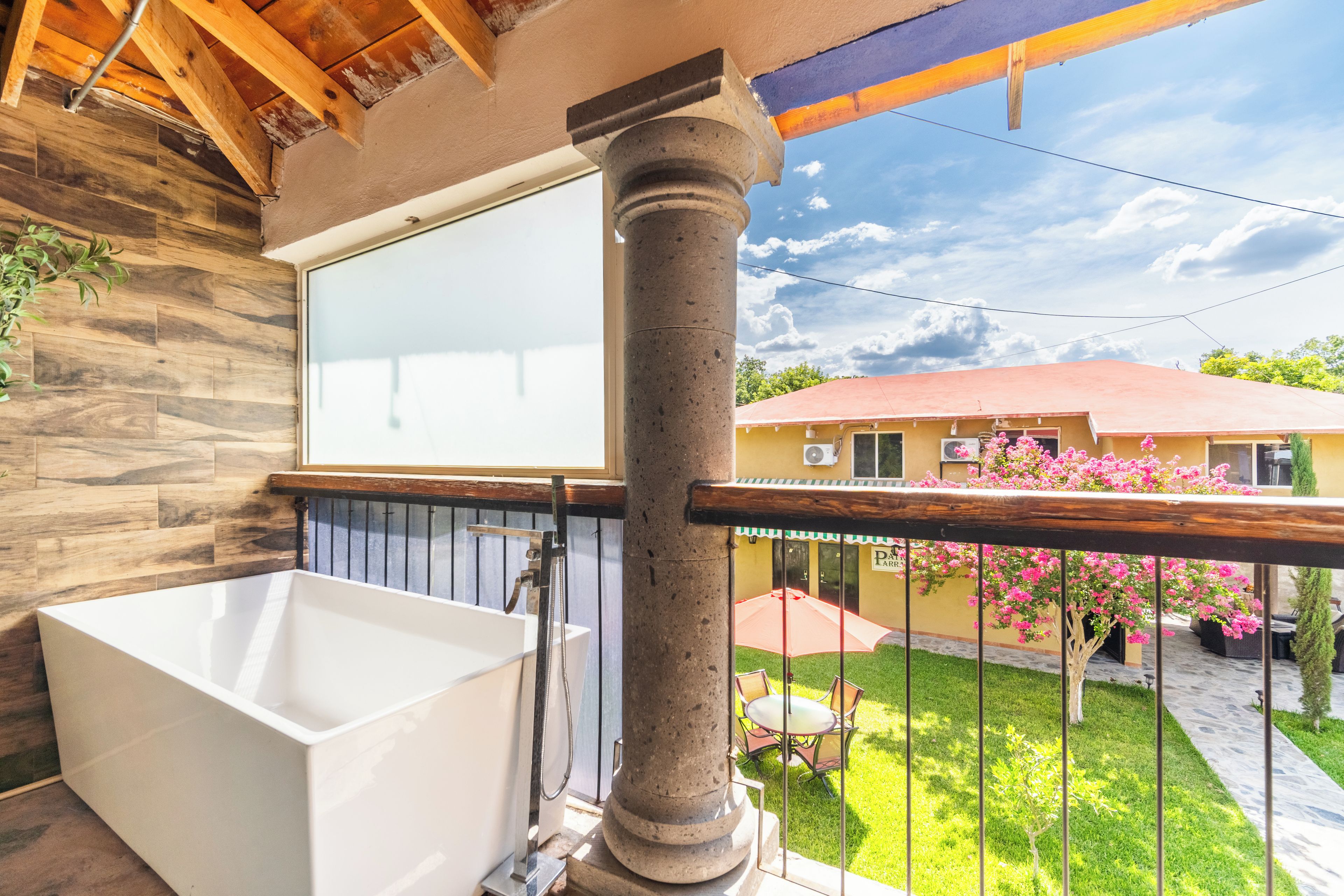 Un baño con bañera, techo de madera y vistas a un jardín con una casa amarilla.