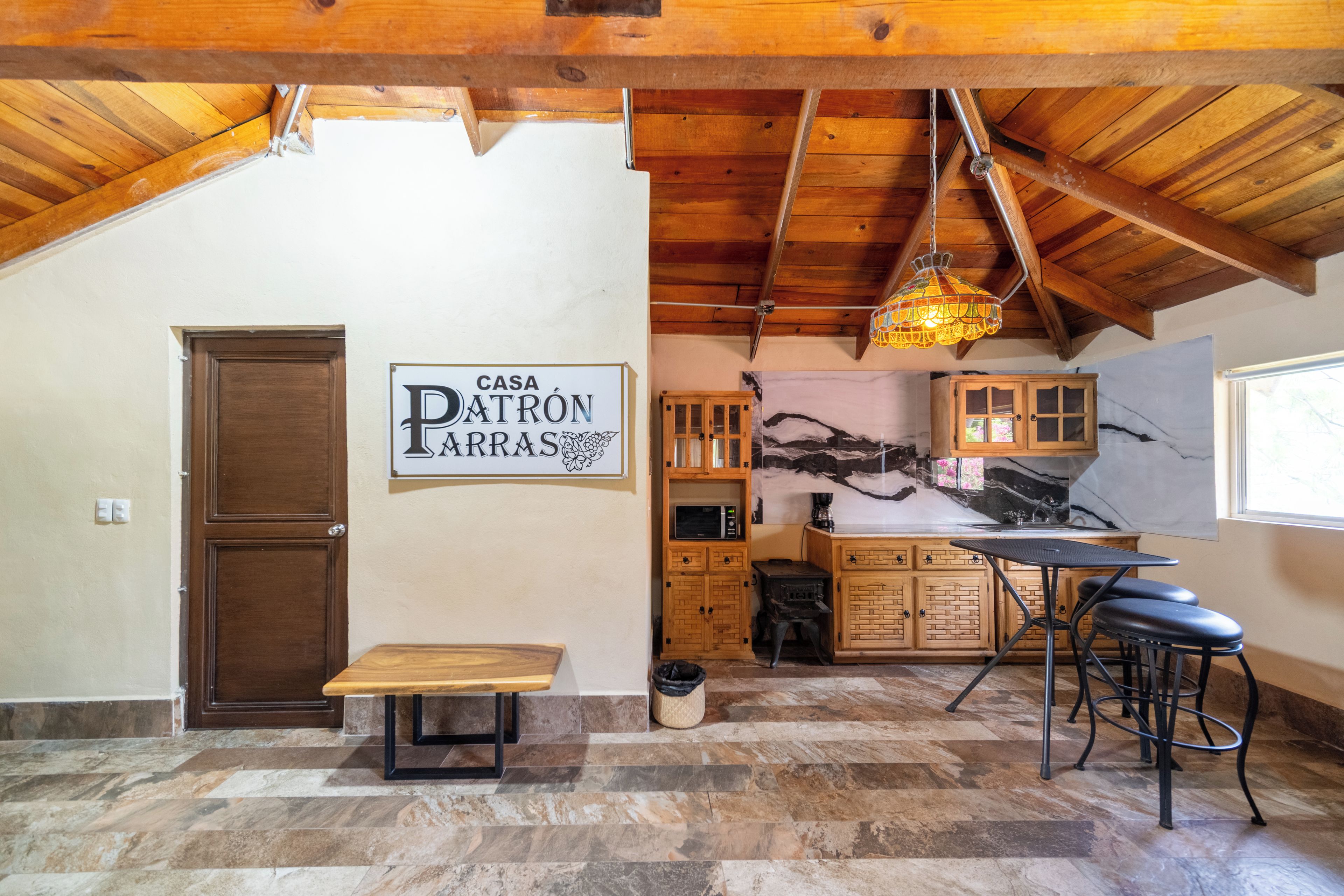 Cozinha rústica com teto de madeira, placa com a inscrição 'CASA PATRON ARRAS', um banco de madeira e uma área de refeições com banquetas.