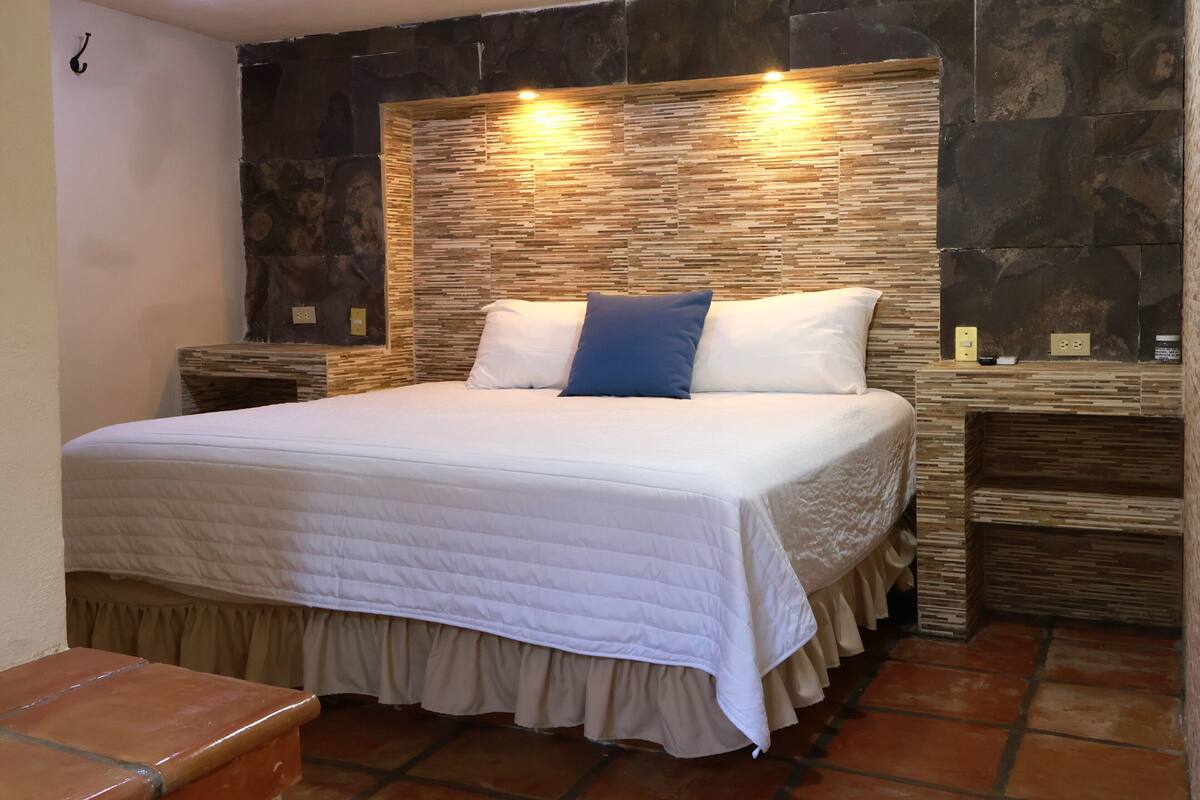A bed with a white quilt, two pillows, a blue accent pillow, and a headboard with a stone-like texture.