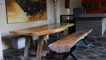 A rustic wooden table with benches, a stone floor, a wall-mounted painting, and a kitchen area.