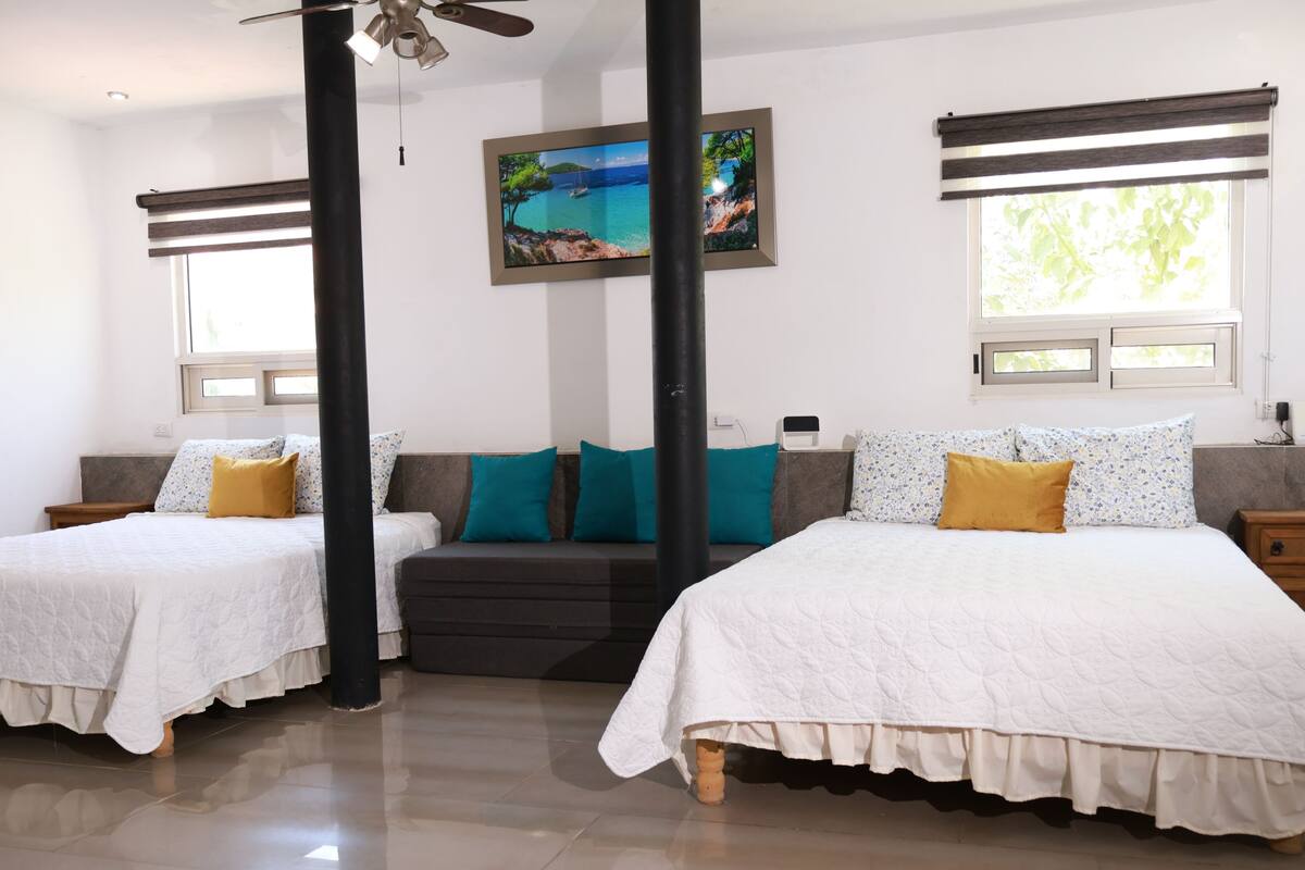 A hotel room with two beds, a sofa, a ceiling fan, a painting, and a window with blinds.
