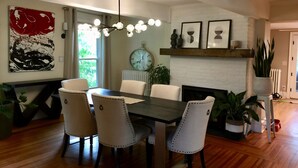 Un coin repas avec une table en bois foncé et des chaises à revêtement blanc. Un grand tableau abstrait orne le mur, et une horloge est fixée au-dessus d’une cheminée.