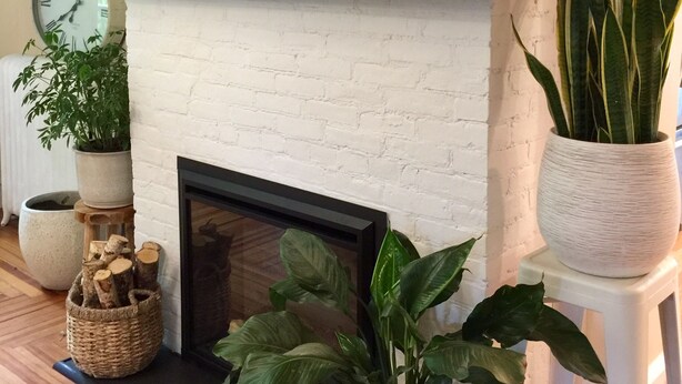 Un salon avec une cheminée en briques blanches, une cheminée en bois ornée de tableaux encadrés, une horloge et diverses plantes en pot.