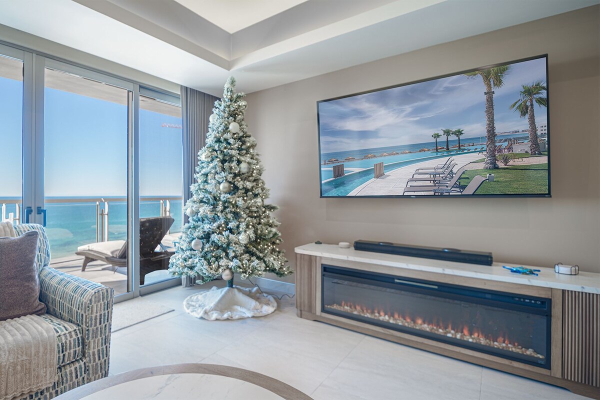 A modern living room with a large window overlooking the sea, a fireplace, a Christmas tree, and a TV displaying a resort view.