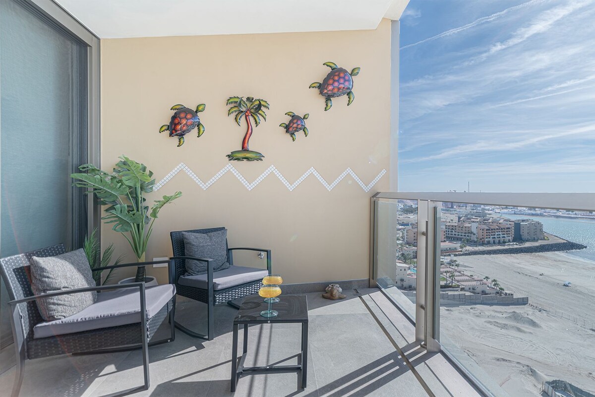 A balcony with a view of the beach, a chair, a table with a drink, and decorative wall art.