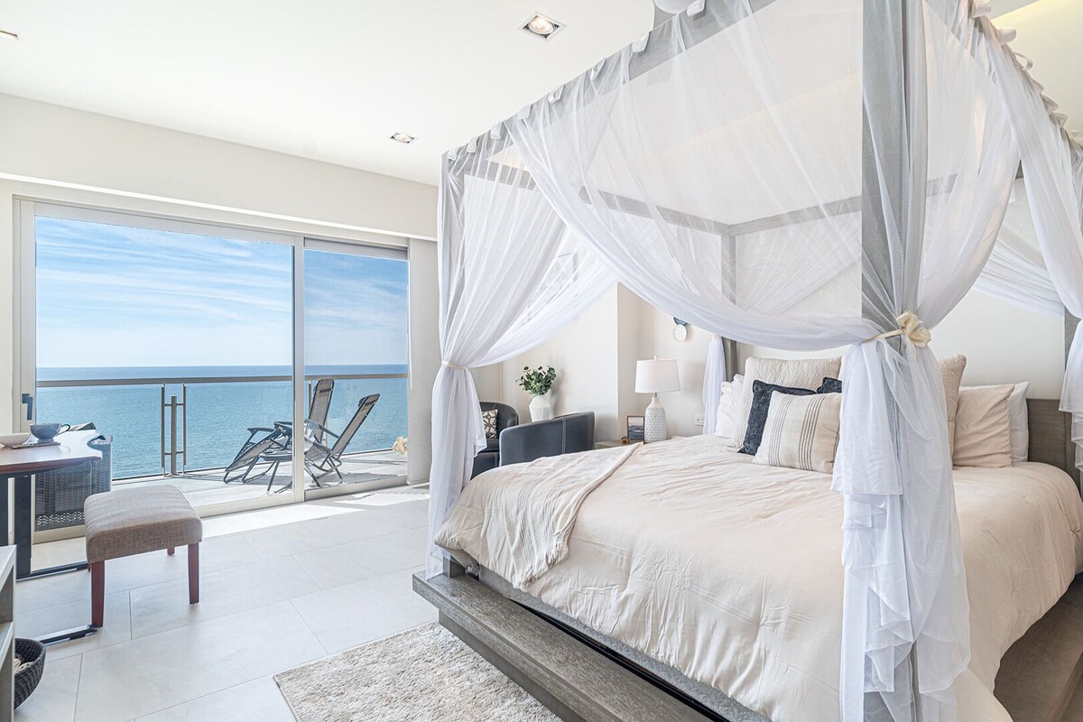 A bedroom with a four-poster canopy bed, a balcony with a view of the sea, and a desk with a chair.