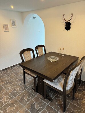 A dining area with a dark wooden table, four chairs with patterned cushions, a decorative wall-mounted deer head, and a stone-tiled floor.