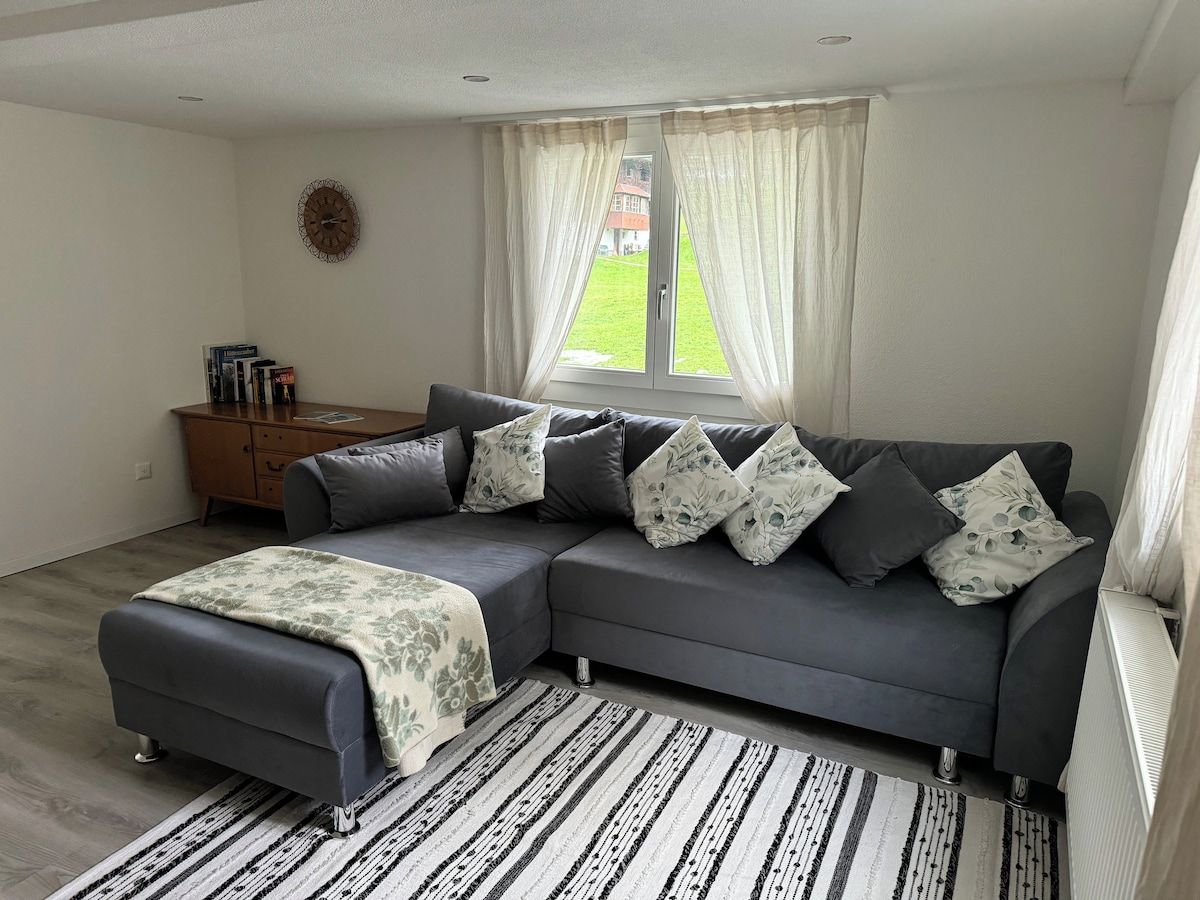 A modern living room with a grey sectional sofa, a patterned throw blanket, a wooden side table, and a view of a green lawn through sheer curtains.