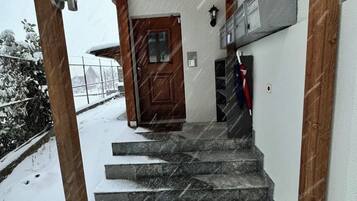 Un escalier menant à une porte en bois, surmontée d’une grille métallique, le tout entouré de neige.