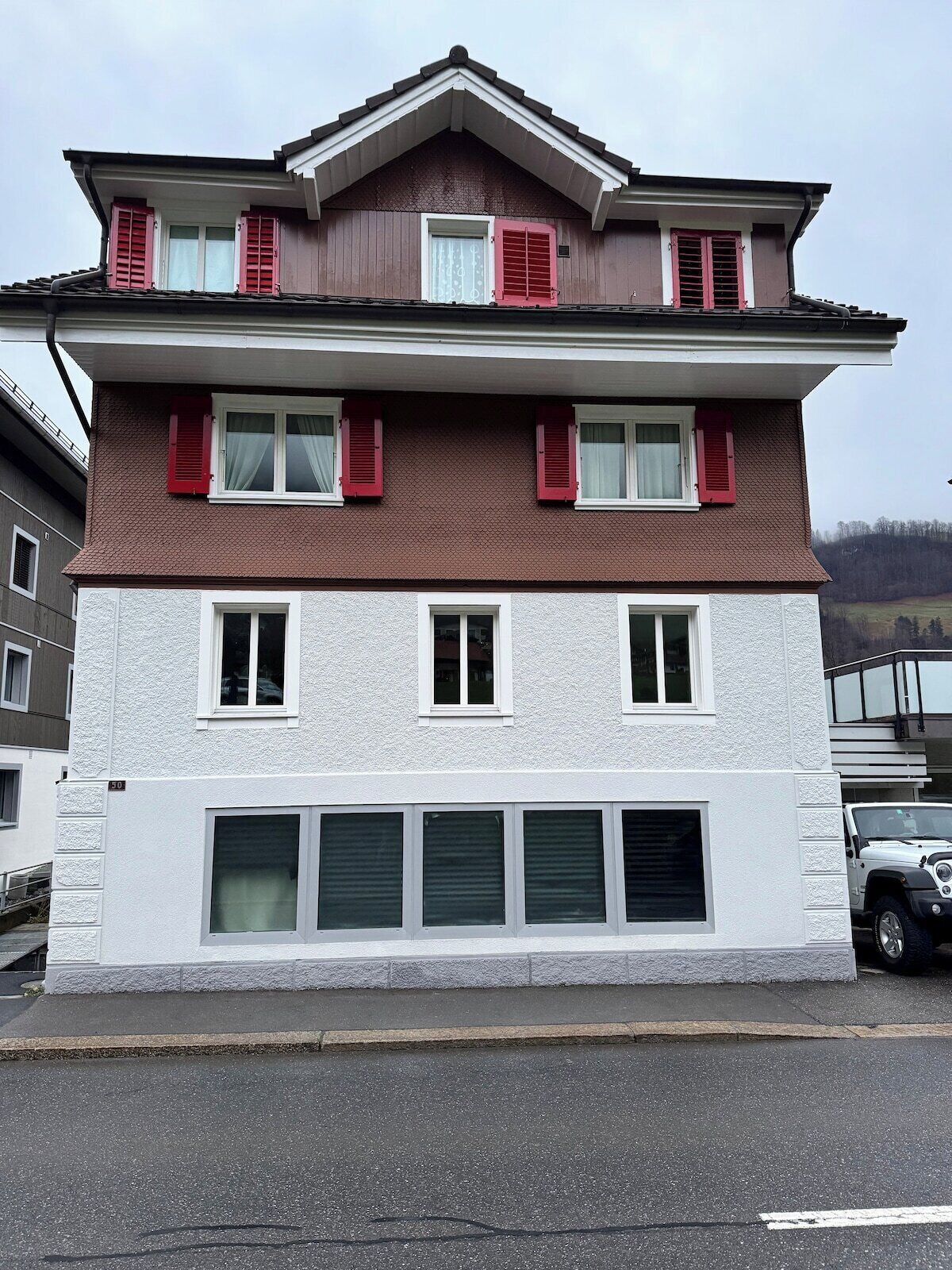 A multi-story building with a flat roof, white exterior walls, and red shutters on the upper windows.