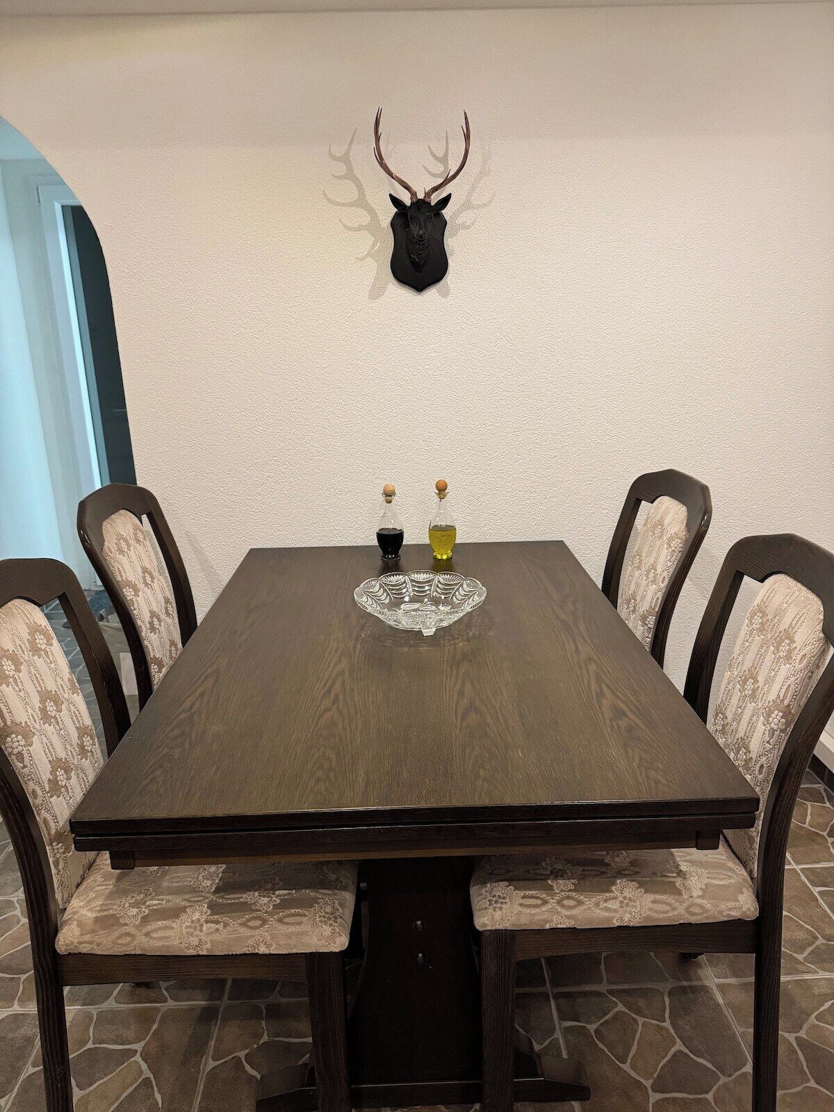 Une table à manger avec six chaises, une tête de cerf décorative fixée au mur et deux bouteilles d’huile d’olive.