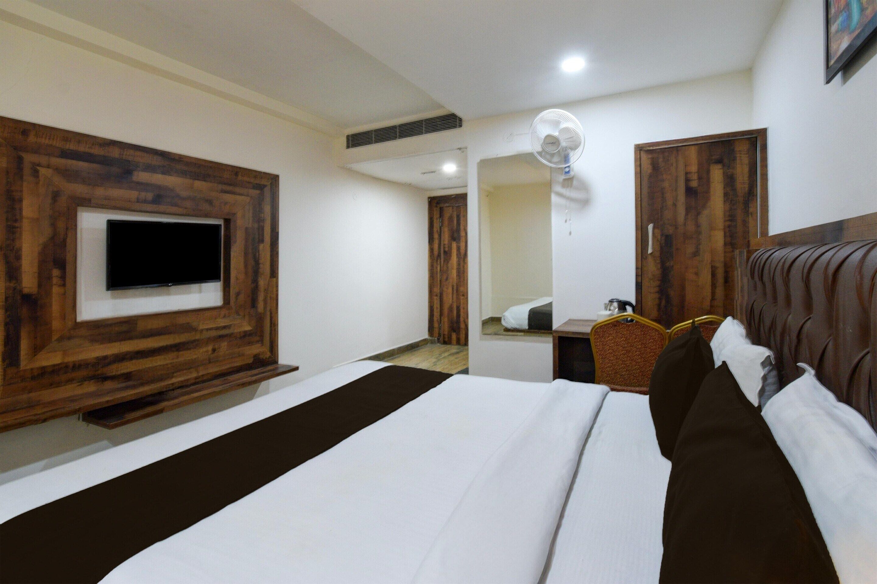 A hotel room with a bed, a television mounted on a wooden panel, a bedside table with a lamp, and a mirror on the wall.