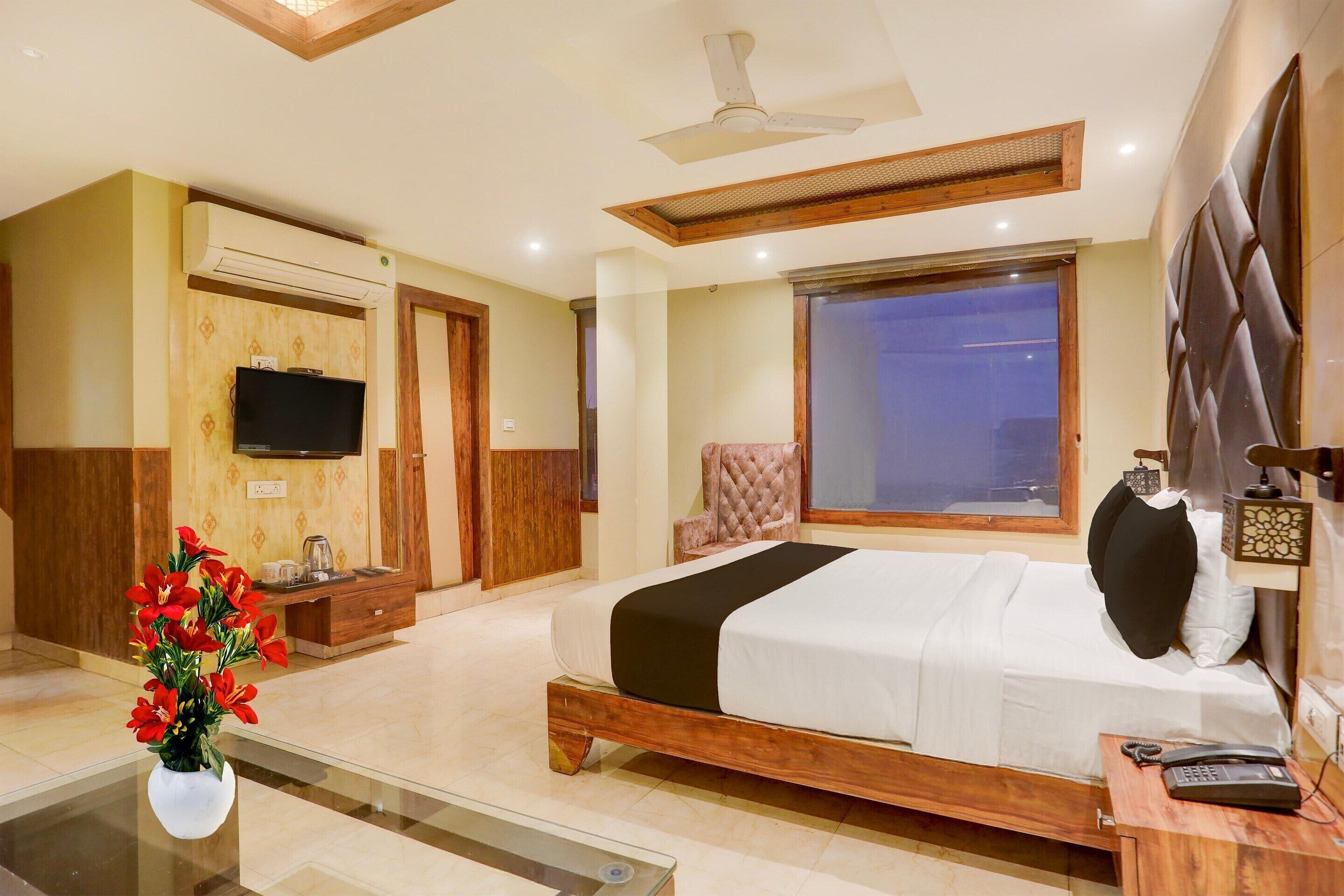 A hotel room with a bed, a TV, a desk with a phone, a vase with flowers, and a window.