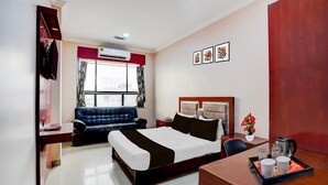 A hotel room with a bed, a desk, a chair, a sofa, a window with curtains, an air conditioning unit, and framed pictures on the wall.