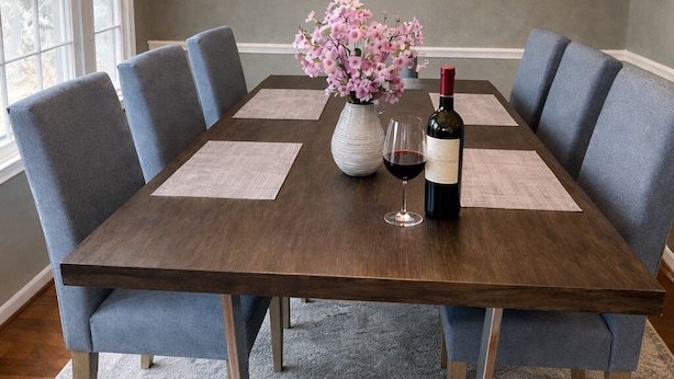 Een eetkamer met een houten tafel, zes blauwe stoelen met bekleding, een vaas met roze bloemen, een wijnglas en een fles wijn.
