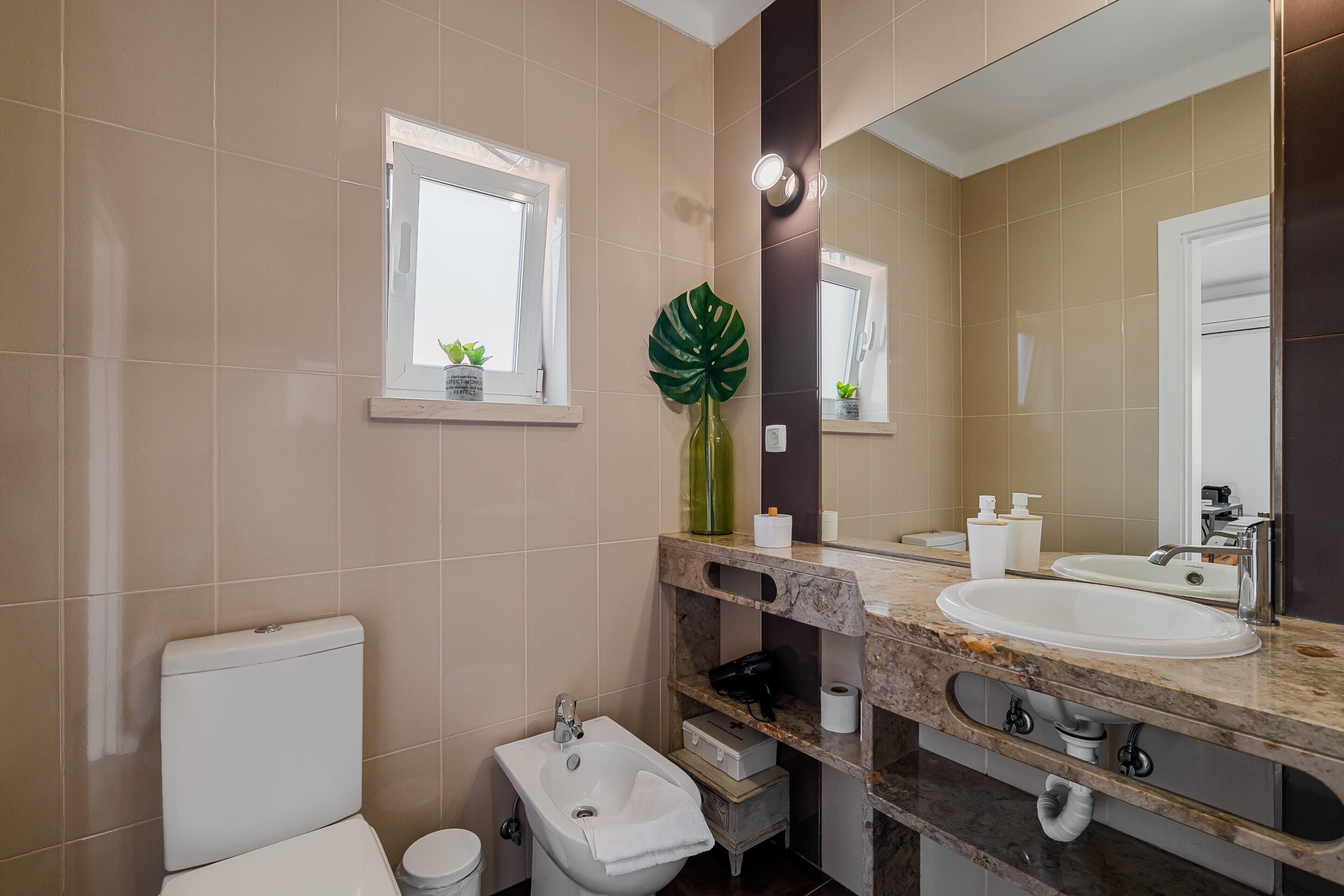A bathroom with a toilet, bidet, sink, mirror, and a window with a plant.