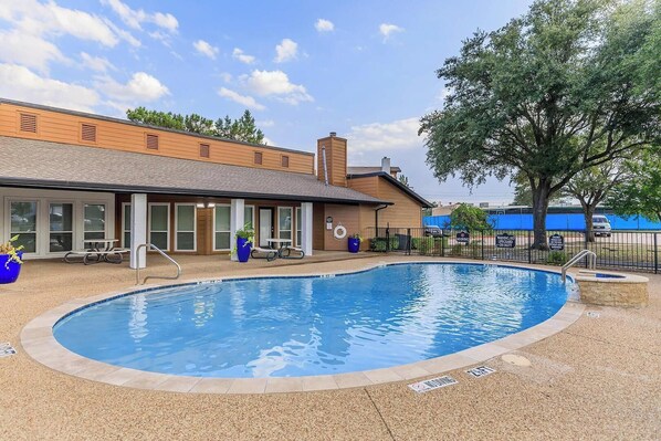 A swimming pool with a tiled deck, a covered patio area with tables and chairs, and a building with large windows and a brick facade. - Landing Apartments Near CVC (DeSoto)