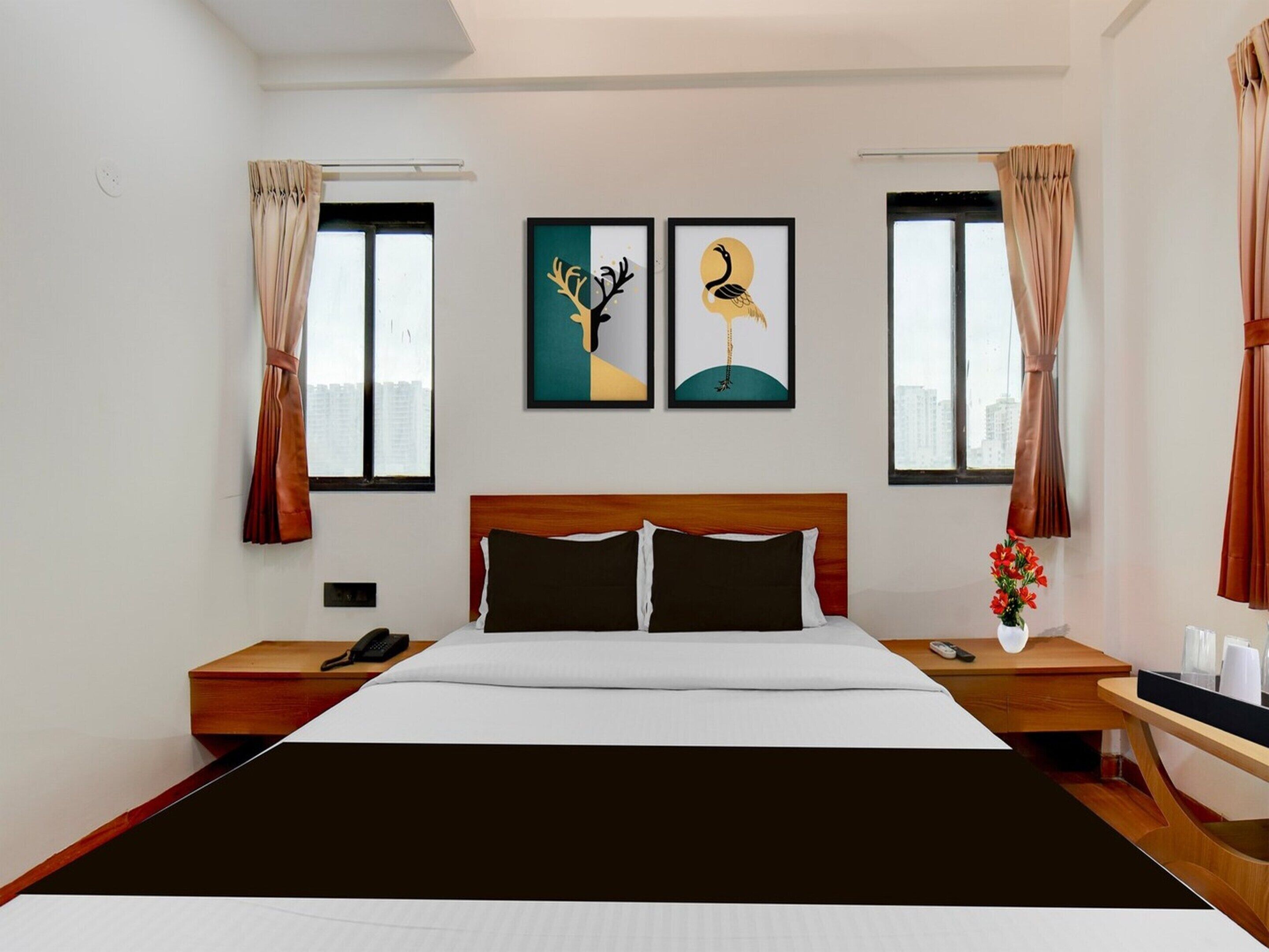 Quarto de hotel com uma cama grande, duas mesas de cabeceira, um criado-mudo, um vaso com flores e duas fotos emolduradas na parede.