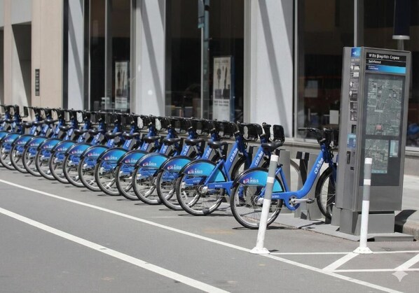 Una fila de bicicletas azules estacionadas frente a un edificio con un panel informativo de mapas.