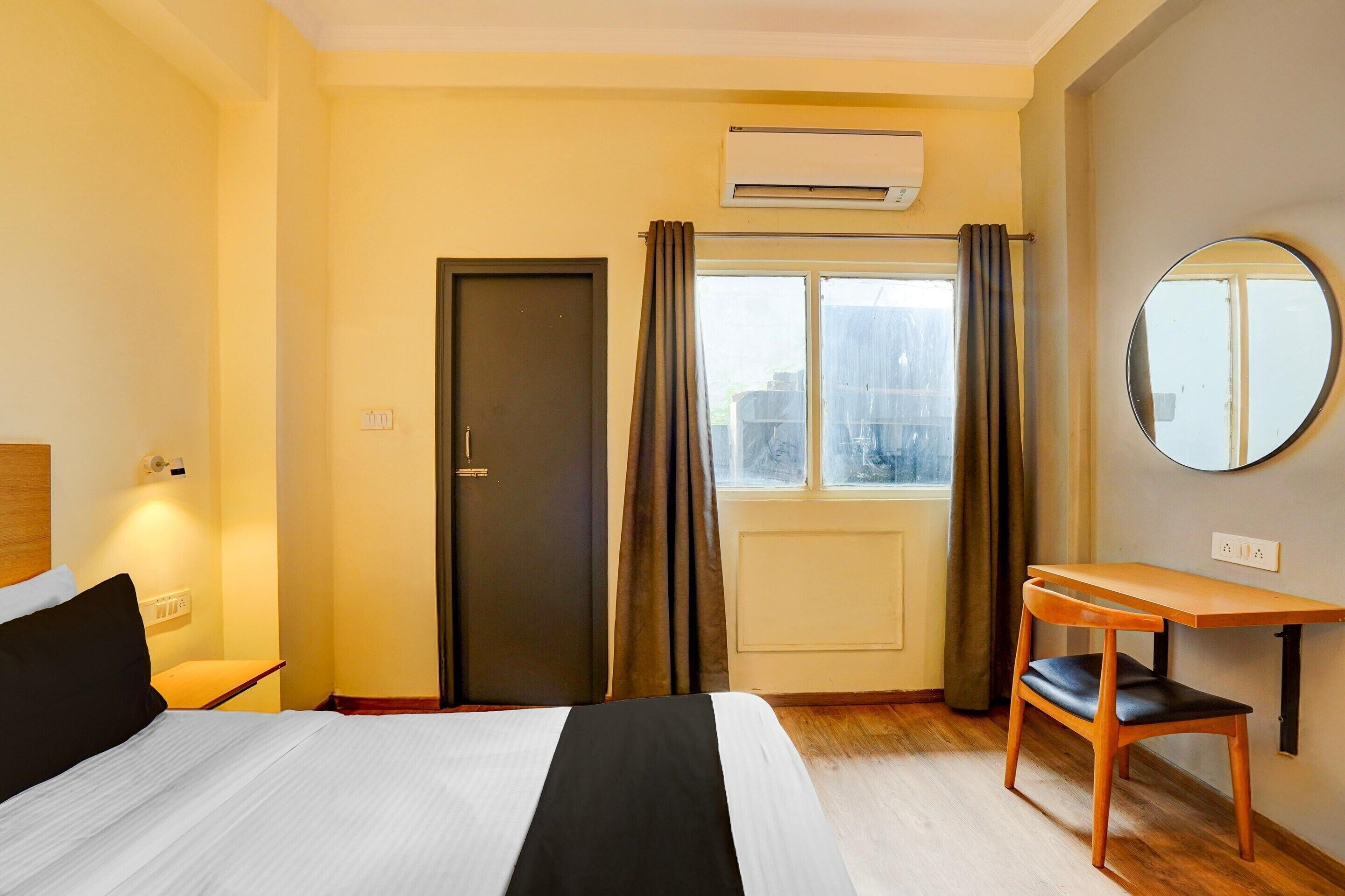 Ein Hotelzimmer mit Bett, Schreibtisch, Stuhl, einem Fenster mit Vorhängen, einer Klimaanlage und einem Spiegel.