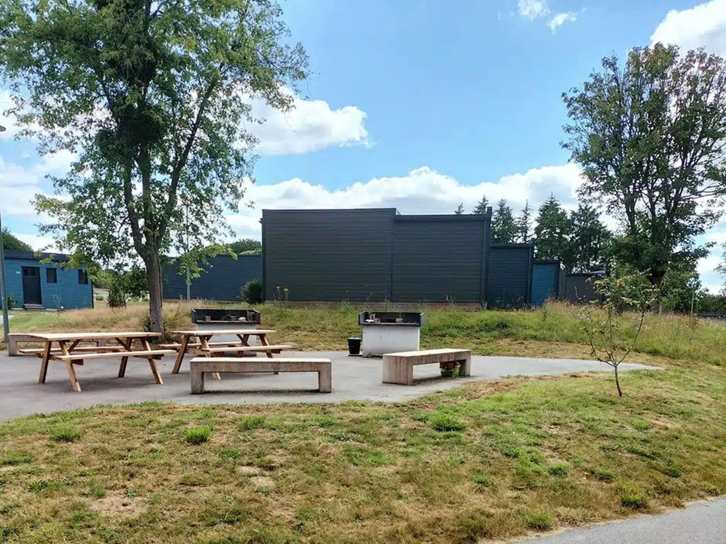 Un espacio exterior con mesas de picnic y bancos, un campo cubierto de césped y un edificio grande al fondo.