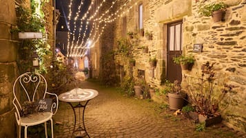 Un edificio de piedra con un patio adoquinado, una silla de metal blanco, una mesa redonda y luces colgantes.