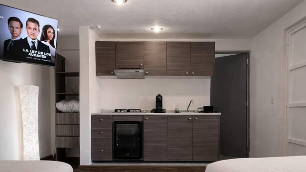 A modern hotel room with a kitchenette, two beds, and a TV displaying a show. - Milan Providencia Studio (Guadalajara)
