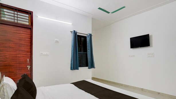 A hotel room with a bed, a TV mounted on the wall, a window with curtains, and a door with a wooden panel. - Deluxebellandur Near Embassy Tech Village (Bakshi Ka Talab)