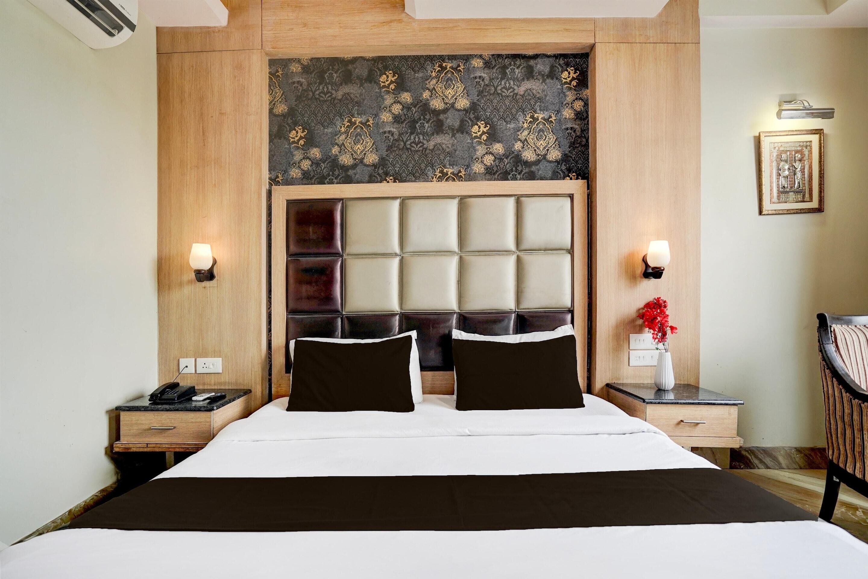 A hotel room with a large bed, wooden headboard, two bedside tables, a nightstand, a wall-mounted lamp, and a framed picture.