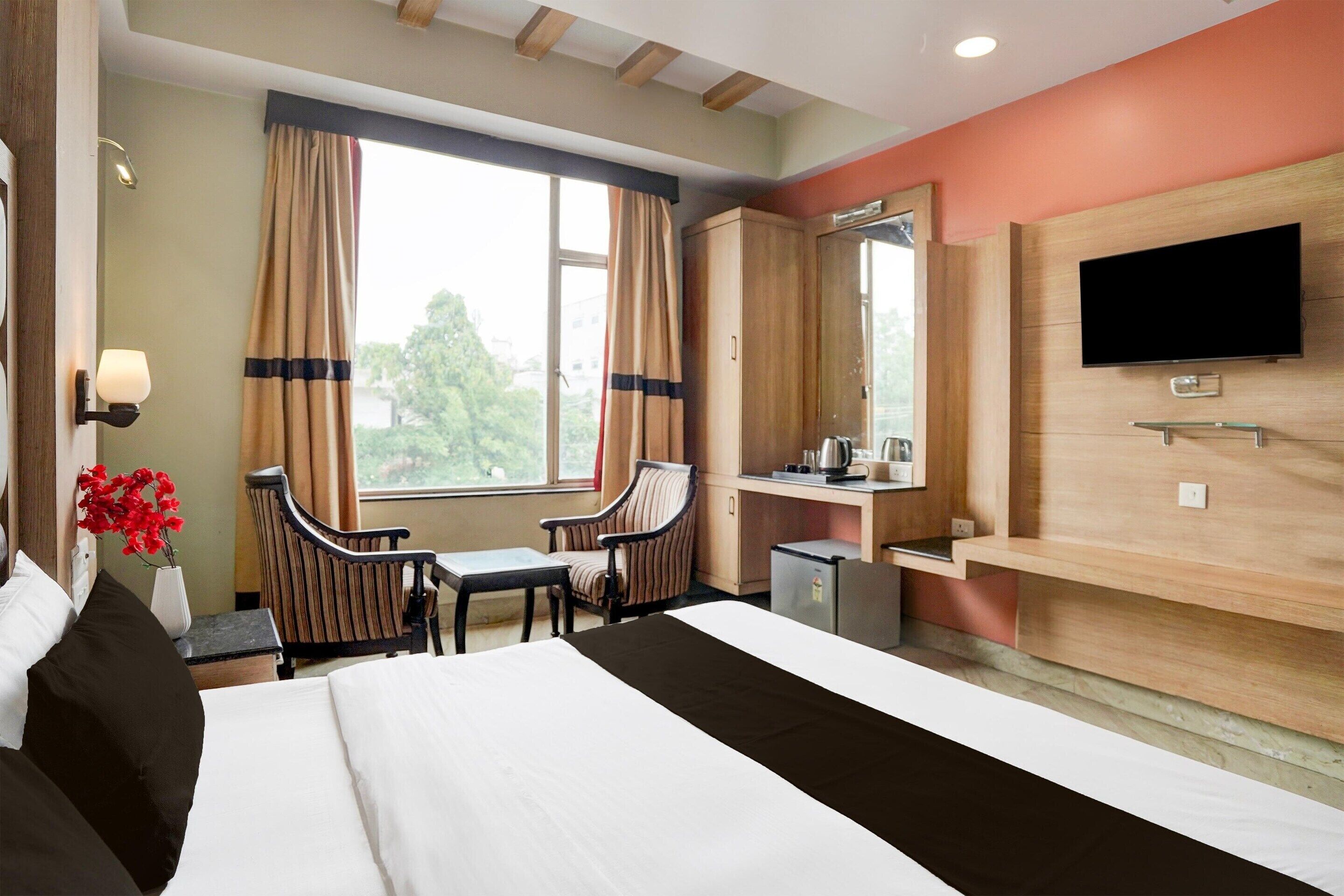 Une chambre d’hôtel avec un lit, deux chaises, un bureau, une télévision et une fenêtre avec des rideaux.