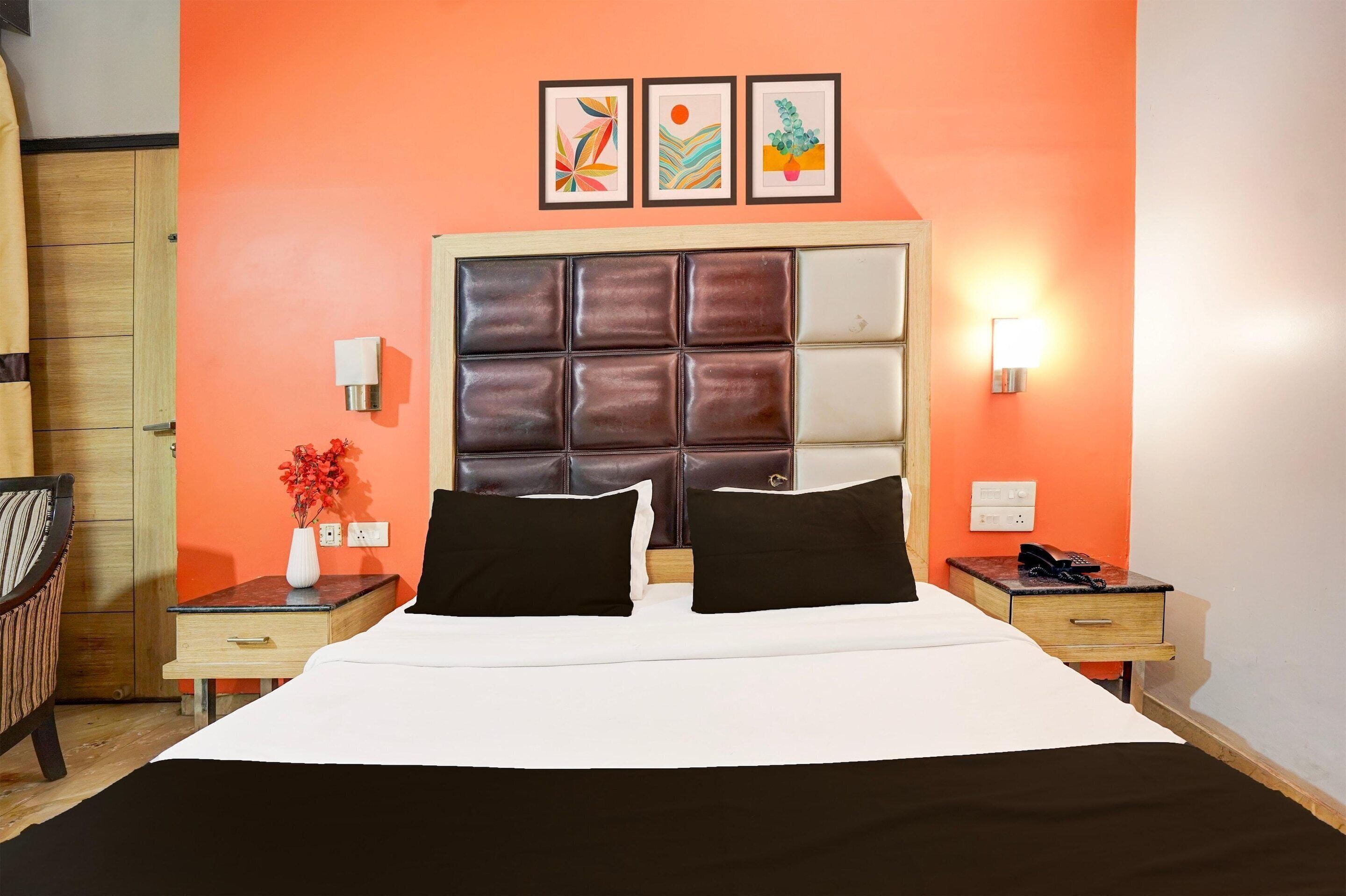 Una habitación de hotel con una cama, una mesita de noche, una lámpara, un florero con flores y una pared con tres cuadros.