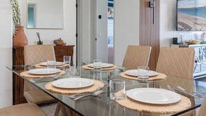 A dining table set with plates, glasses, and cutlery, surrounded by cushioned chairs, with a view of the beach through the window.
