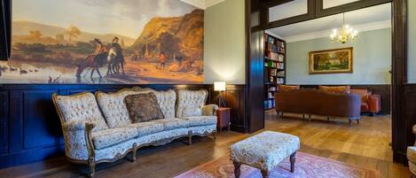 A room with a large mural, a sofa, an ottoman, a bookshelf, and a chandelier.