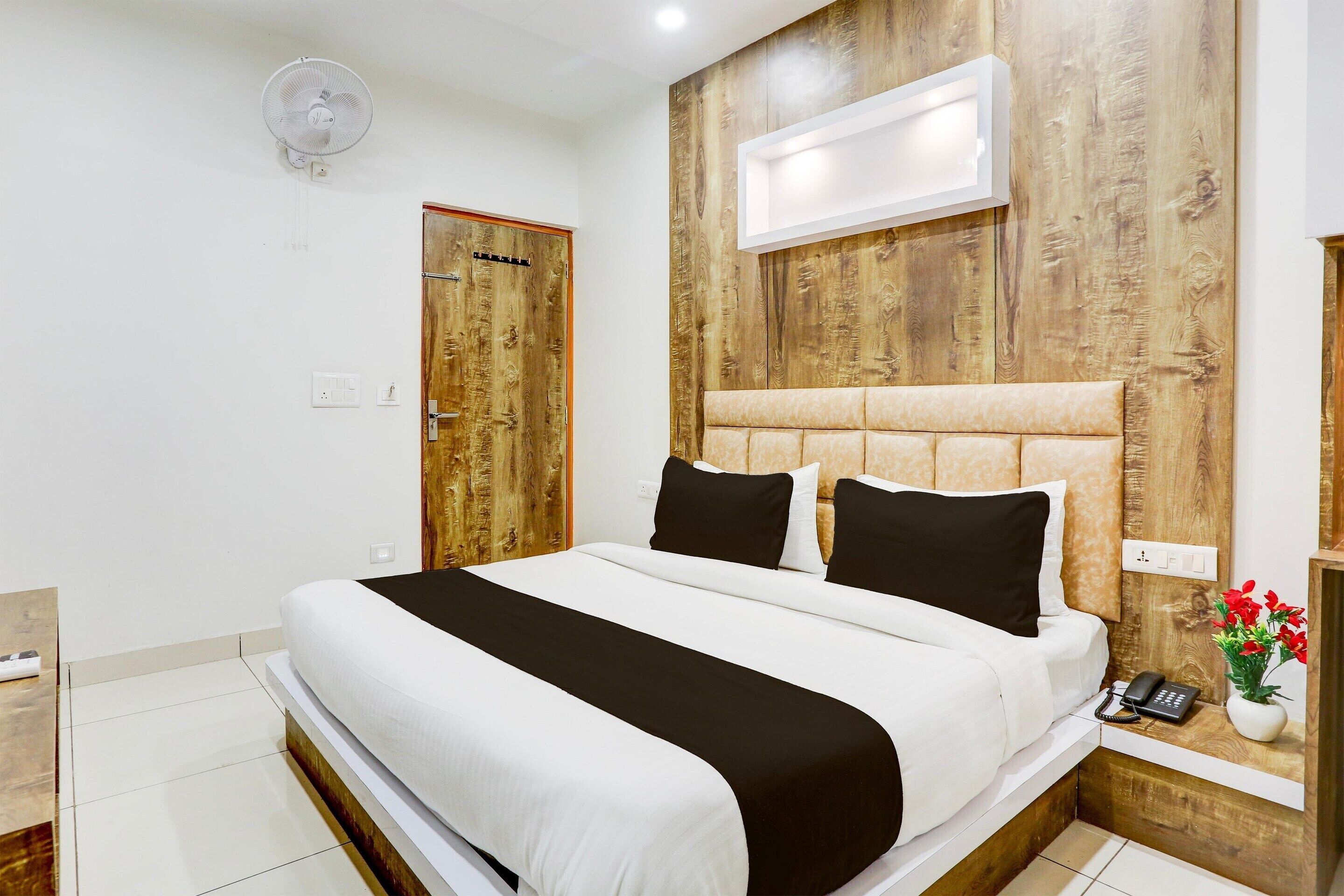 A hotel room with a bed, bedside table, fan, and a wall-mounted air conditioning unit.